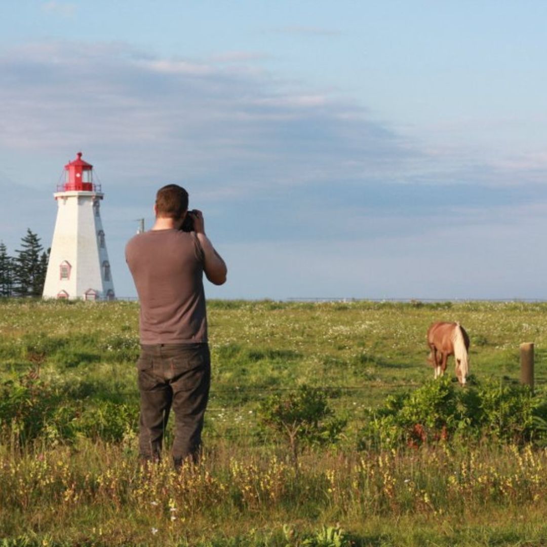 Boston-based photographer Aaron Spagnolo stands in a field, photographing a lighthouse and a horse in coastal Canada.