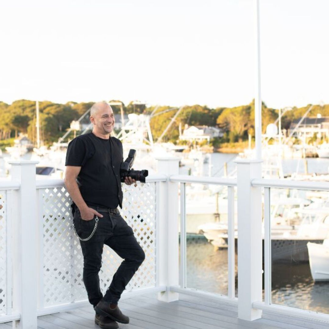 Boston wedding photographer Aaron Spagnolo leans against a white fence on the patio of the Wychmere Beach Club during a wedding the Spagnolo team was photographing.