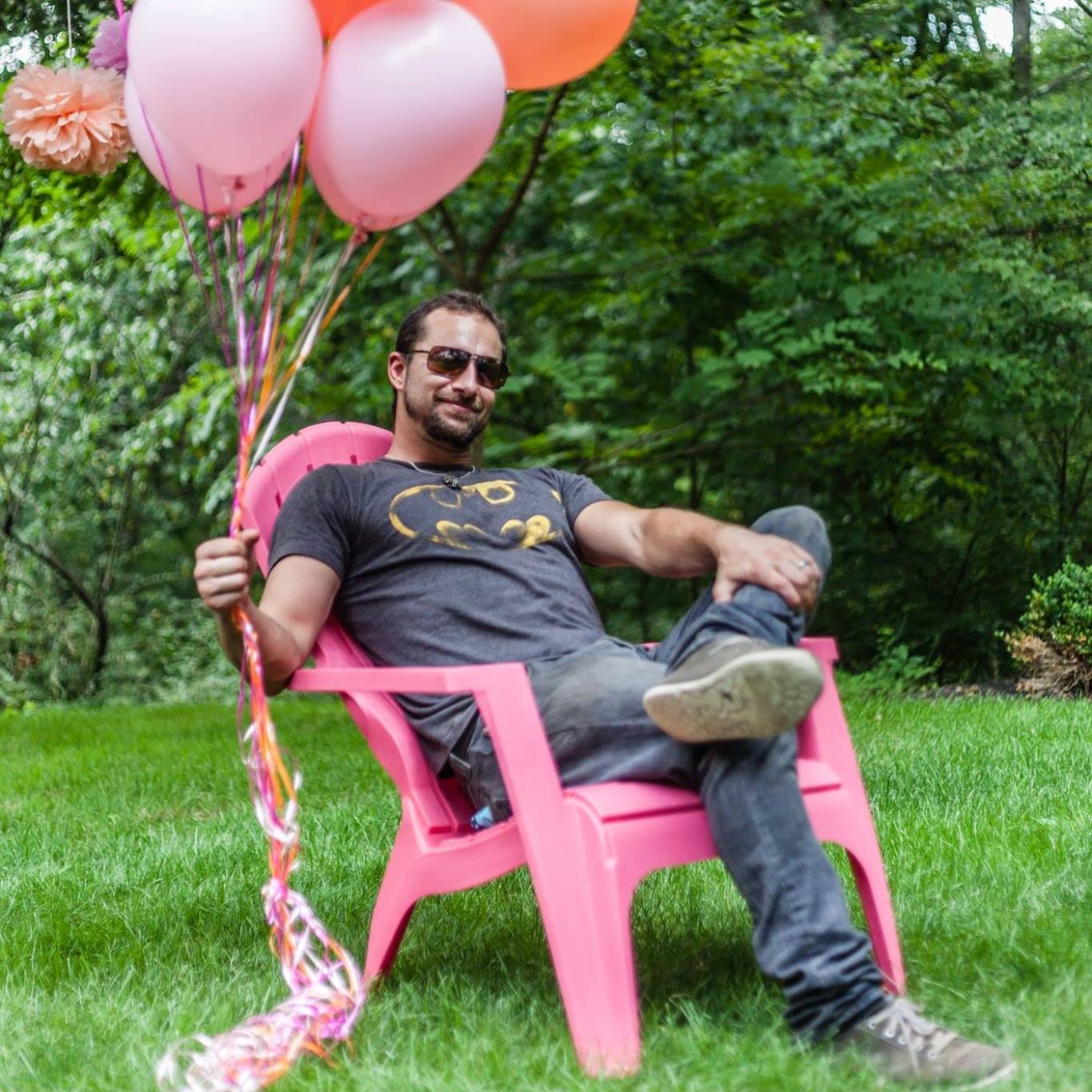 Boston wedding photographer Aaron Spagnolo of Spagnolo Photography wearing a Batman T-shirt, holding a bunch of pink balloons, and sitting in a pink chair.