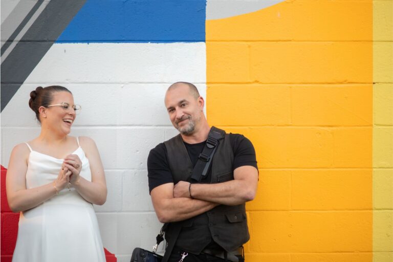 Aaron leans against a yellow wall to demonstrate a pose for the bride standing and laughing next to him during a late summer wedding at Night Shift Brewery in Everett, MA.