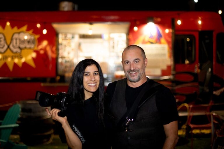 Boston wedding photographers Aaron and Nanore of Spagnolo Photography stand in front of a red food truck under string lights during a wedding they were photographing at Night Shift Brewing in Everett, MA - the photograph, taken by a wedding guest is featured on their "About Us" page.