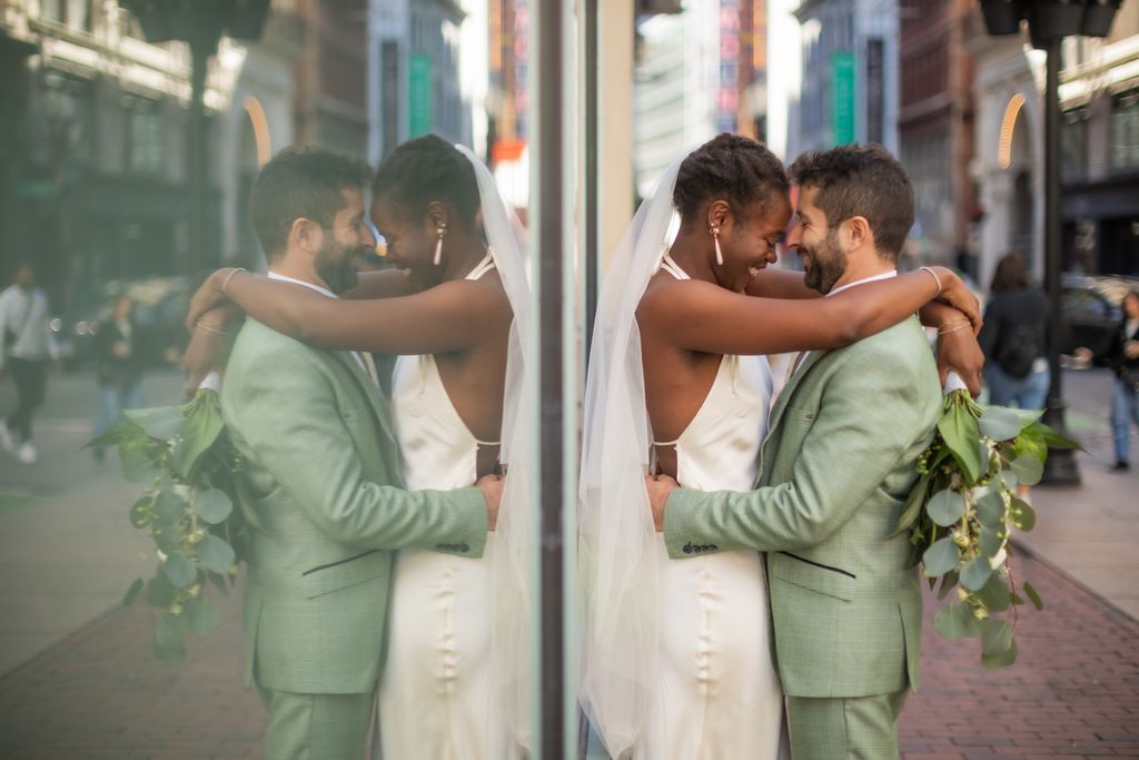 A bride in a wedding gown wraps her arms around the groom's neck, who's wearing a light green wedding suit, foreheads touching, smiles on their faces - and their reflections are mirrored in the glass wall behind them - the photograph is part of their Boston elopement photography package by Spagnolo Photography.