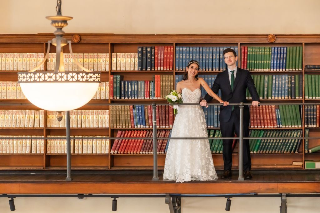 A bride and a groom pose in front of a large bookcase after their Boston Public Library 1-hour ceremony, photographed by their Boston elopement photographer Spagnolo Photography as part of their elopement photography package.