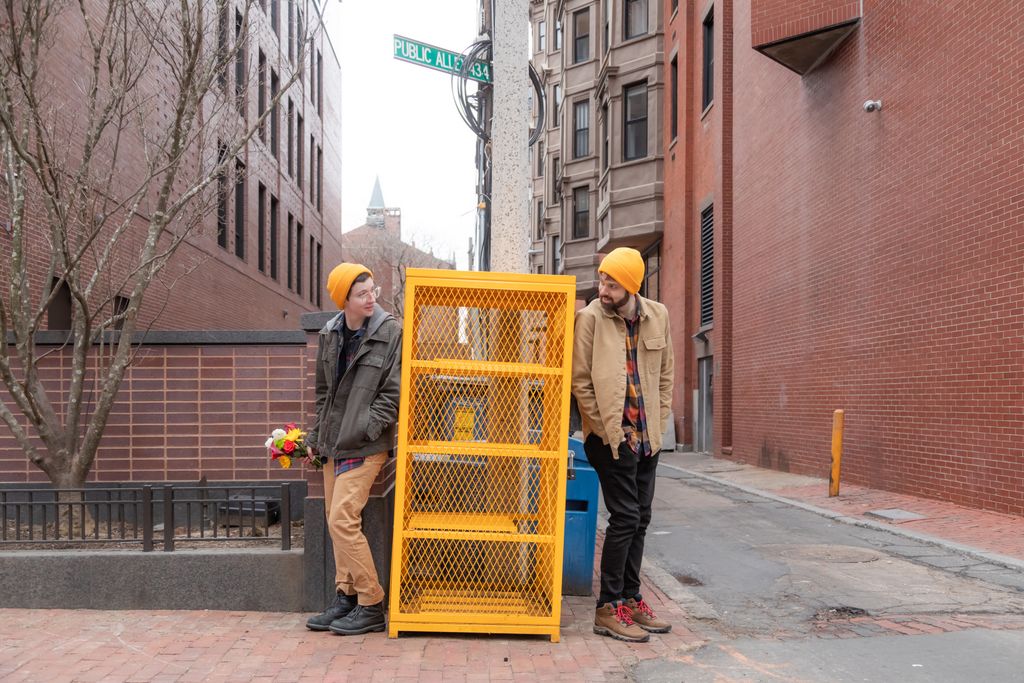 A non-traditional bride and groom, dressed in everyday clothes, both wearing yellow wool hats, stand on either side of an abandoned yellow metal shelf as tall as them, a sunflower bouquet in the bride's hand - photographed on a Boston street by Boston elopement photographer Spagnolo Photography, who specializes in creative and original wedding portraits (image featuured on their elopement photography packages page)