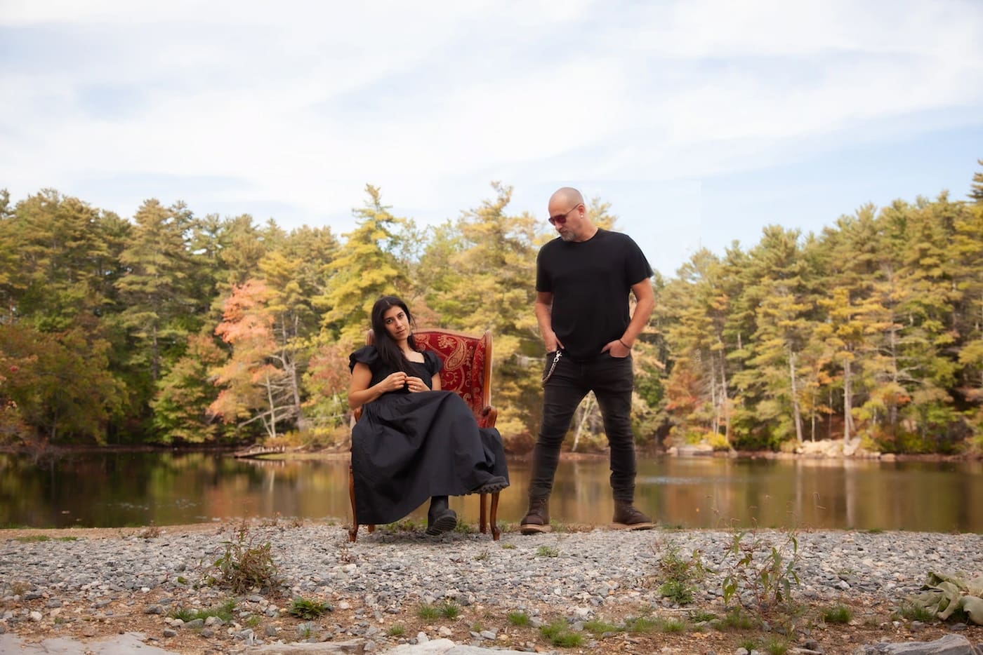 A self-portrait of creative Boston wedding photographers Aaron and Nanore of Sagnolo Photography, showing Nanore dressed in a black dress and sitting in a red armchair at the edge of a lake and Aaron in black pants and a black shirt standing next to her, looking at her, fall foliage behind them on the trees - the photograph is featured on their "About Us" page.