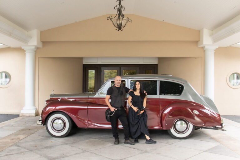 Aaron and Nanore of Spagnolo Photography posing in front of a red Rolls Royce during a wedding they photographed