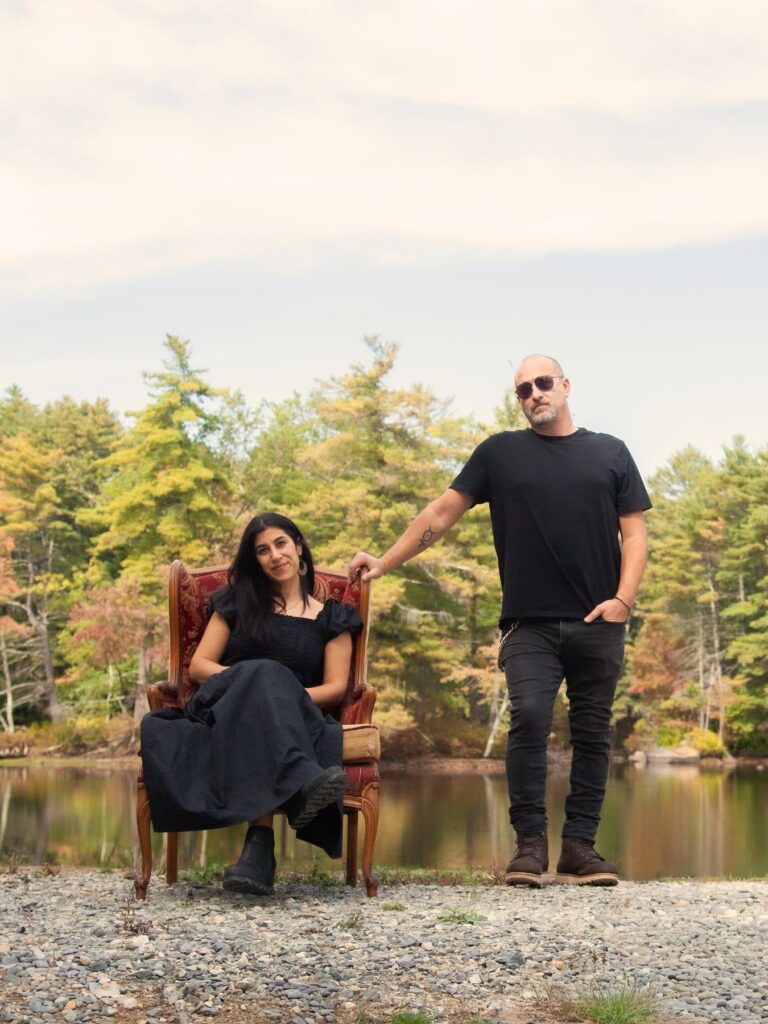 A fun self-portrait of Boston wedding photographers Aaron and Nanore, showing Nanore sitting in a red armchair and Aaron standing next to her in a scenic park with a lake behind them and trees changing color - the photograph is featured on their website