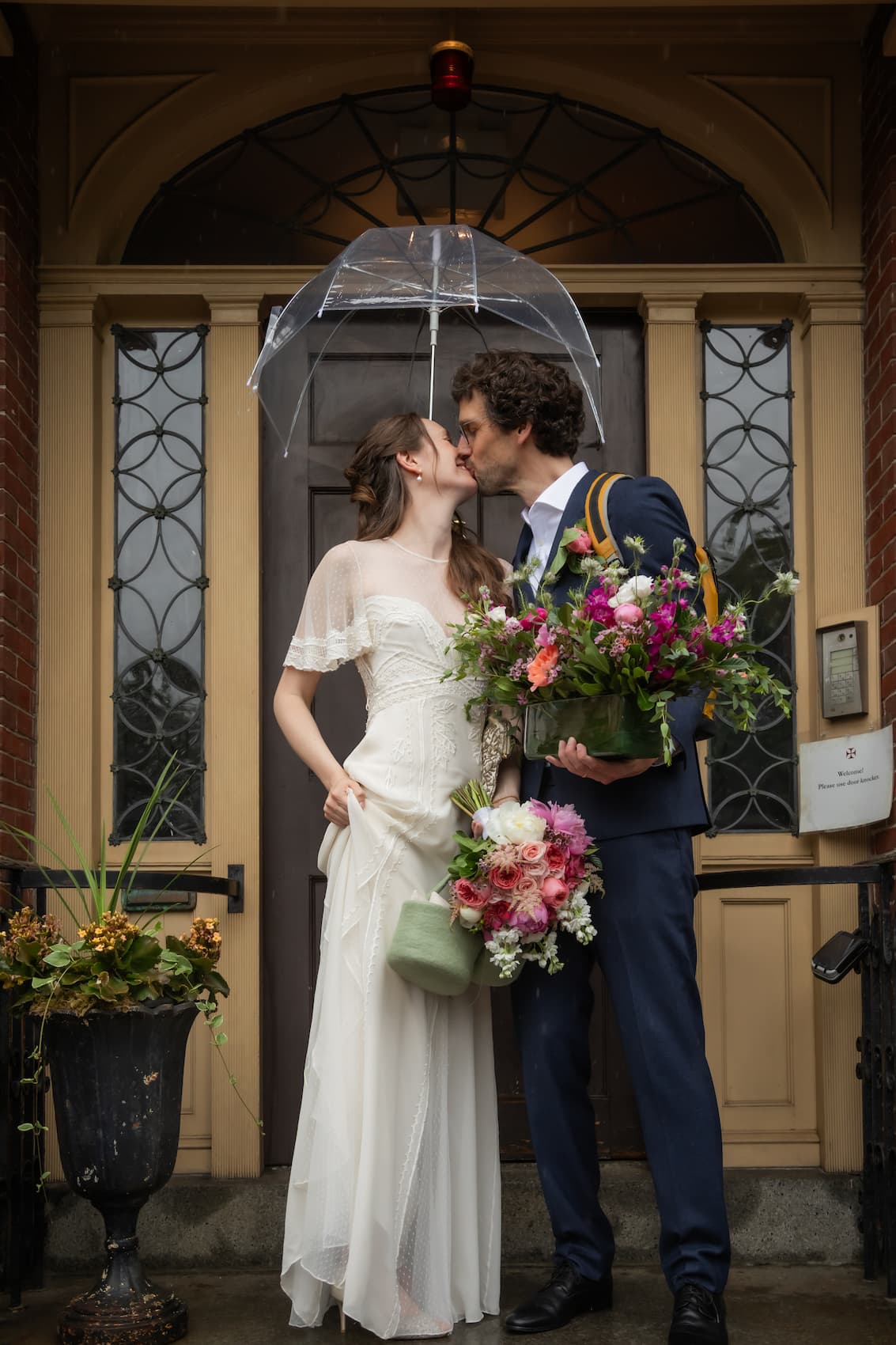 A bride and a groom carrying flowers kiss under a clear umbrella in front of the yellow doorway of the King's Chapel Parish House in Boston's Beacon Hill neighborhood on their wedding day, photographed by Boston wedding and micro-wedding photographers Spagnolo Photography.