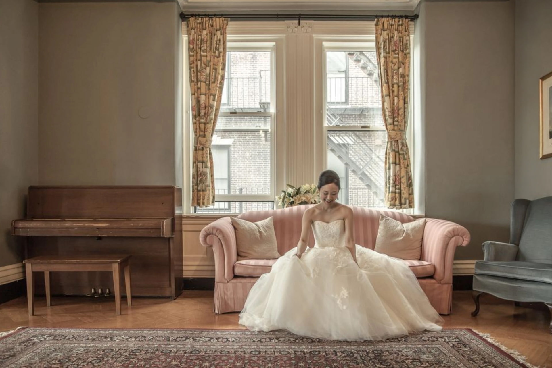 A bride sits on a pink couch and admires her dress in a room with a piano and gray walls at the Boston Union Club during her wedding, photographed by creative Boston wedding photographers Spagnolo Photography as part of her wedding photography package.