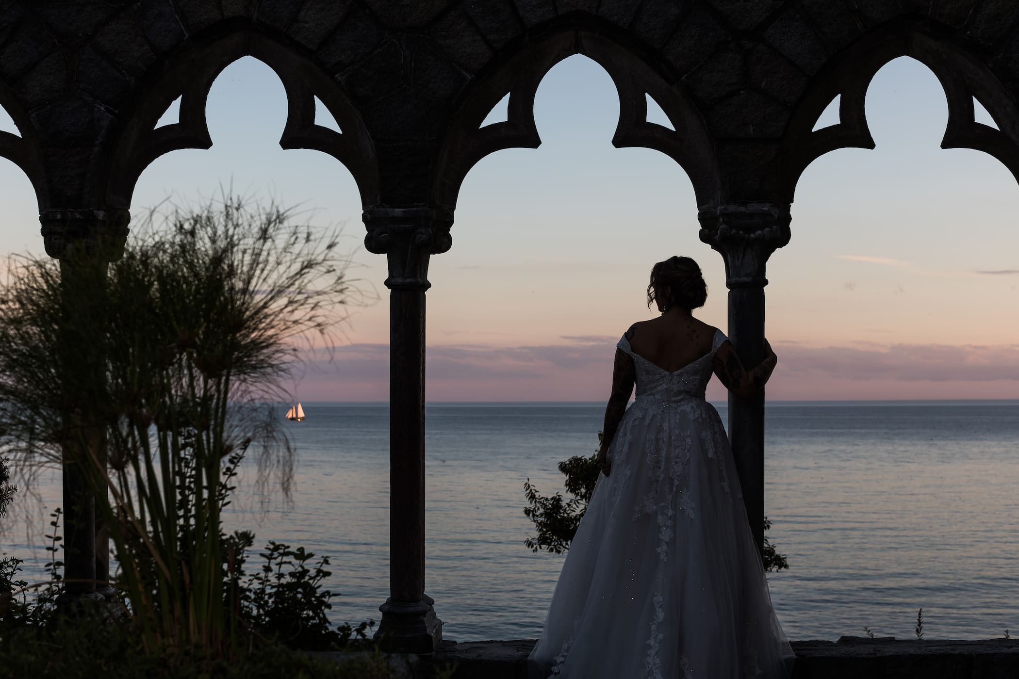 A bride looking out at the ocean and a sailboat at the Hammond Castle arches at sunset, photographed by Massachusetts wedding photographers Spagnolo Photography during her Hammond Castle wedding in Gloucester, MA.
