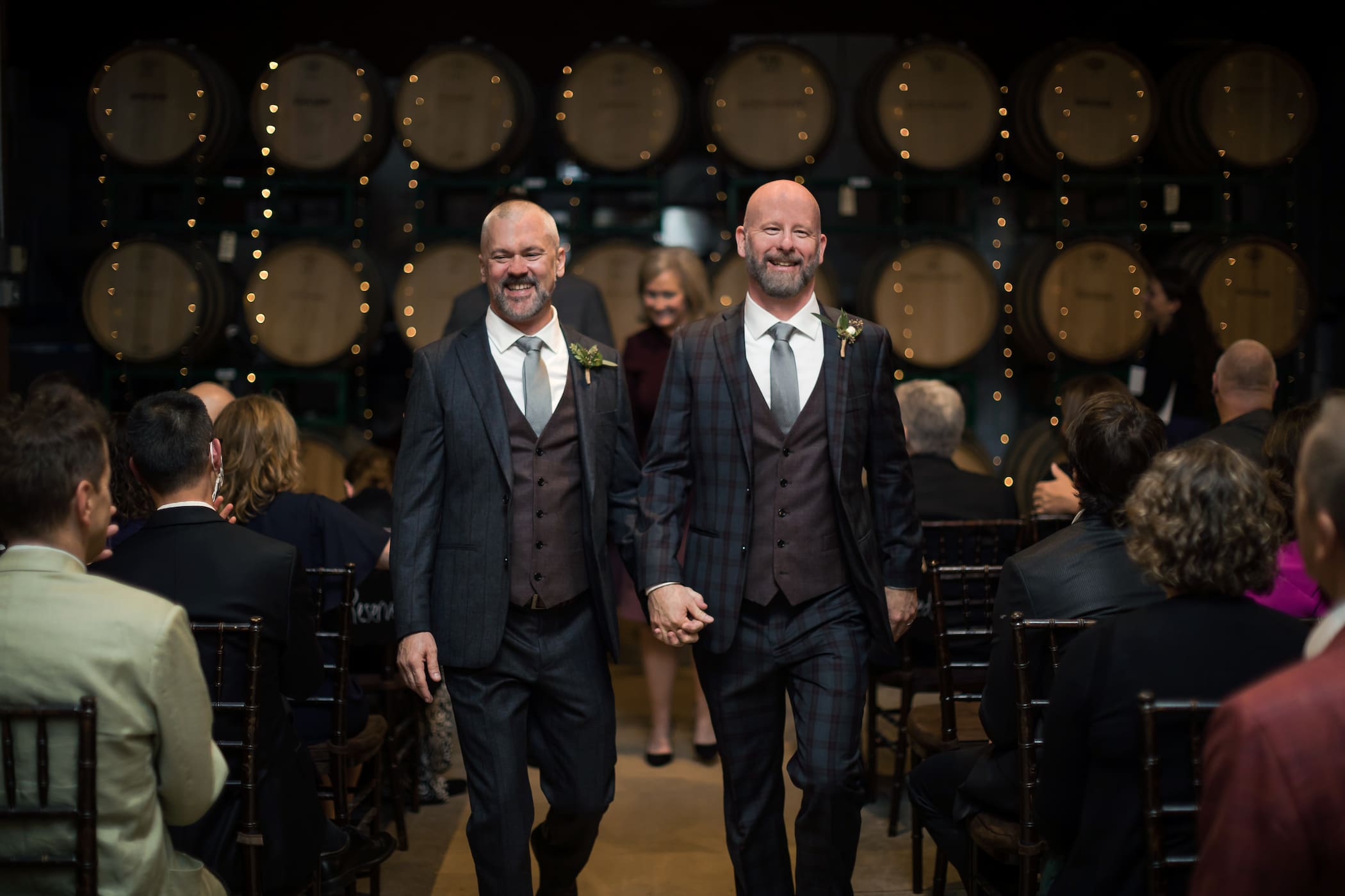 Two grooms in charcoal gray suits, one with a plaid pattern, walk away from the altar holding hands, wooden wine barrels behind them, at a gay wedding at the Boston Winery, photographed by Boston-based LGBTQ friendly wedding photographers Spagnolo Photography.