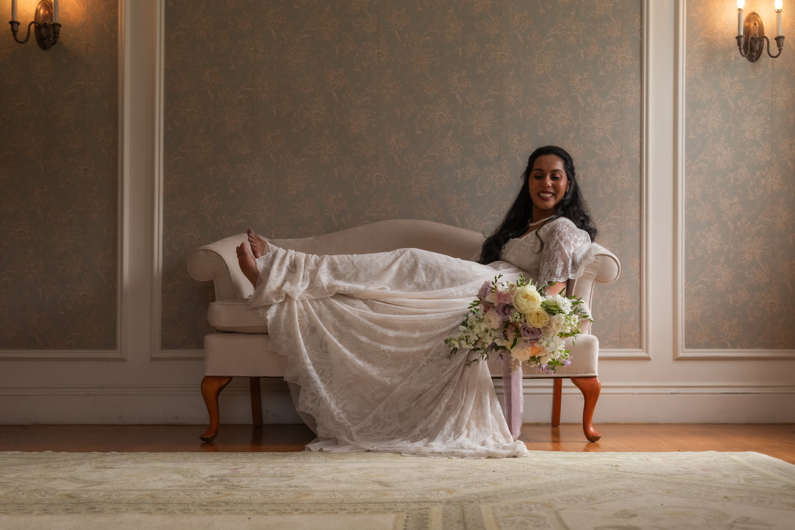A bride lounges on a vintage cream-colored couch, holding a large bouquet of flowers, before the start of her wedding ceremony at the Endicott Estate in Dedham, MA - photographed by Spagnolo Photography as part of her Boston wedding photography package that also included an engagement session.