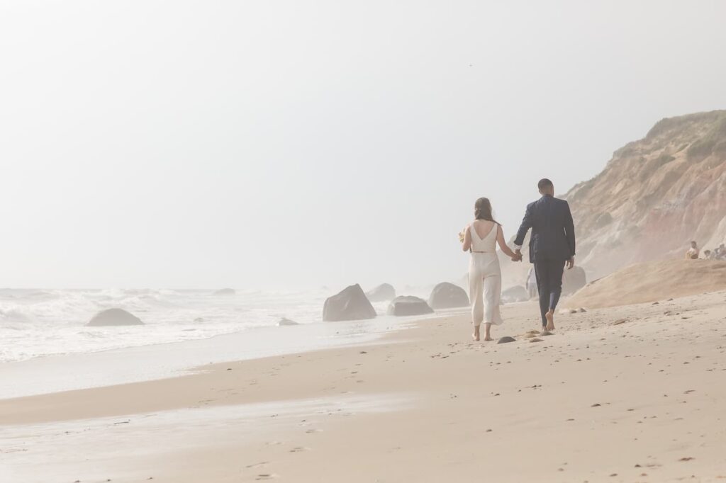 A hazy image of a bride and groom - groom in a navy suit and bride in a white sleeveless jumpsuit - walking away from the camera on the beach holding hands - photo featured in a glowing review of the photographer's work by the bride