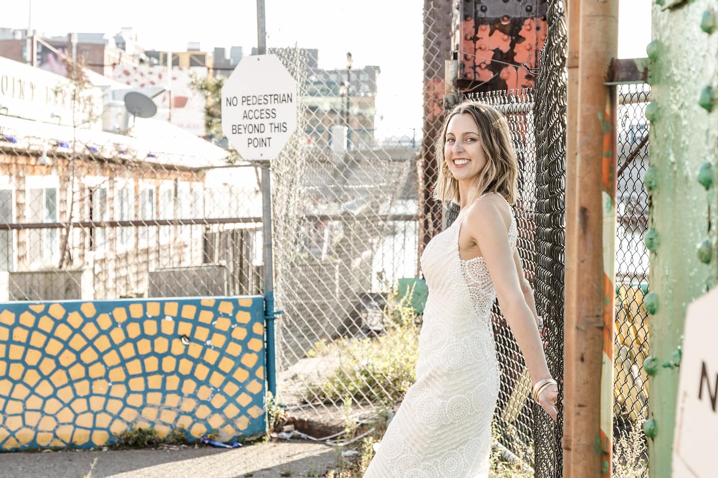 A bride smiles at the camera while leaning against a chain link fence in Boston's Seaport area, before boarding the Odyssey cruise ship for her Boston micro-wedding, photographed by Spagnolo Photography as part of her micro-wedding photography package.