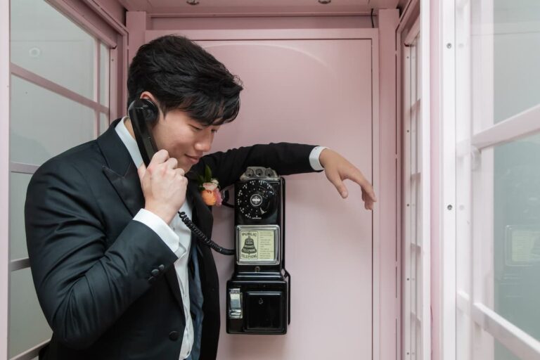 A groom holds the receiver to his ear inside the Langham Boston’s pink phonebooth, captured by Spagnolo Photography and featured on their "Contact Spagnolo Photography" page.
