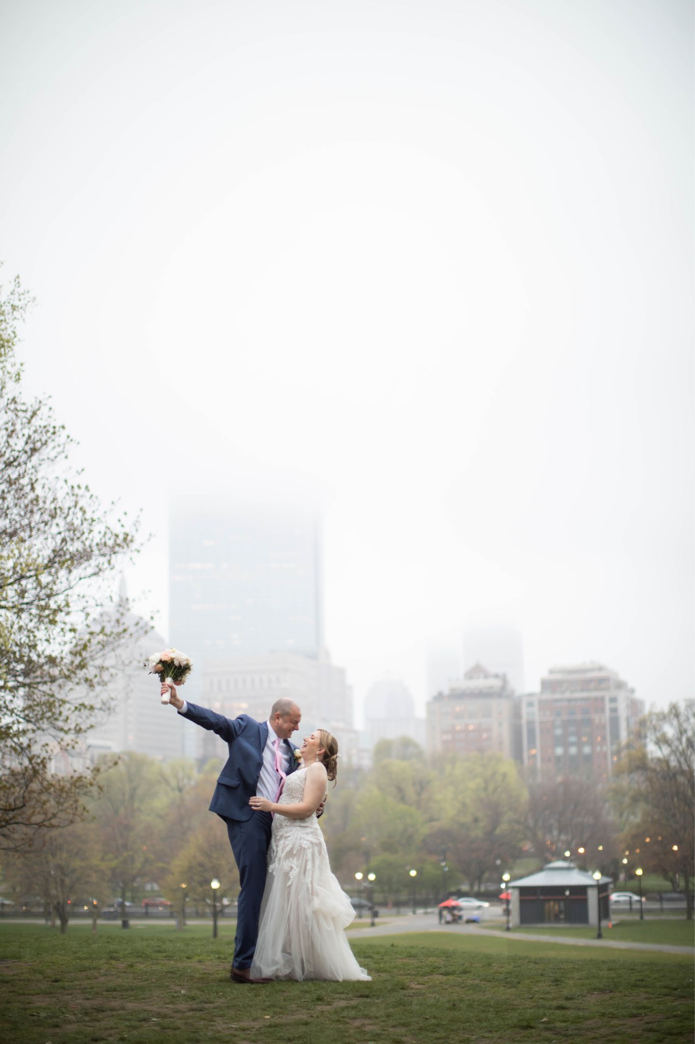 A wedding portrait on the Boston Common of a groom embracing the bride with one arm and with the other raising a flower bouquet, behind them fog envelops the city, photographed during their micro-wedding at the Hampshire House by Spagnolo Photography.