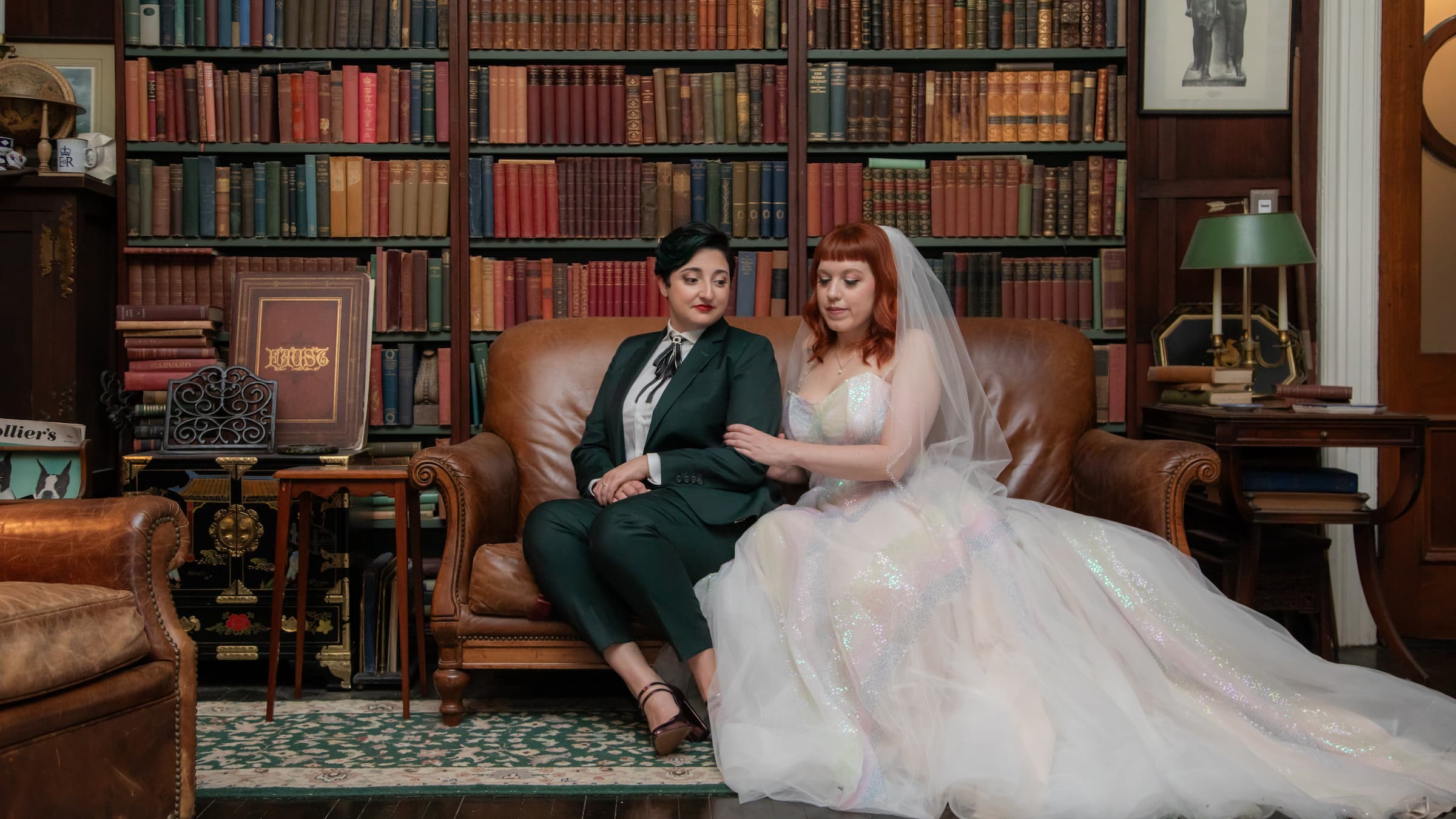 Two brides sit on a brown leather couch surrounded by books and antiques at Diehl Marcus in Salem, MA, before their LGBTQ wedding at The Ledger restaurant, photographed by artistic LGBTQ-friendly wedding photographers Spagnolo Photography.