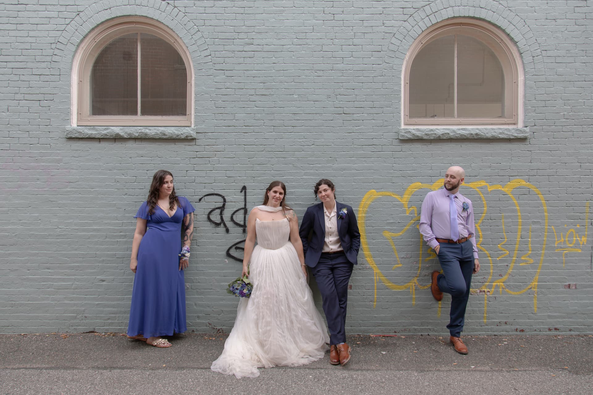 A couple and their bridesmaid and best man lean against a blue-gray wall with graffiti in an alley in Salem, MA, on their way to the Ledger Restaurant for their LGBTQ micro-wedding - photographed by Spagnolo Photography as part of their micro-wedding photography package.
