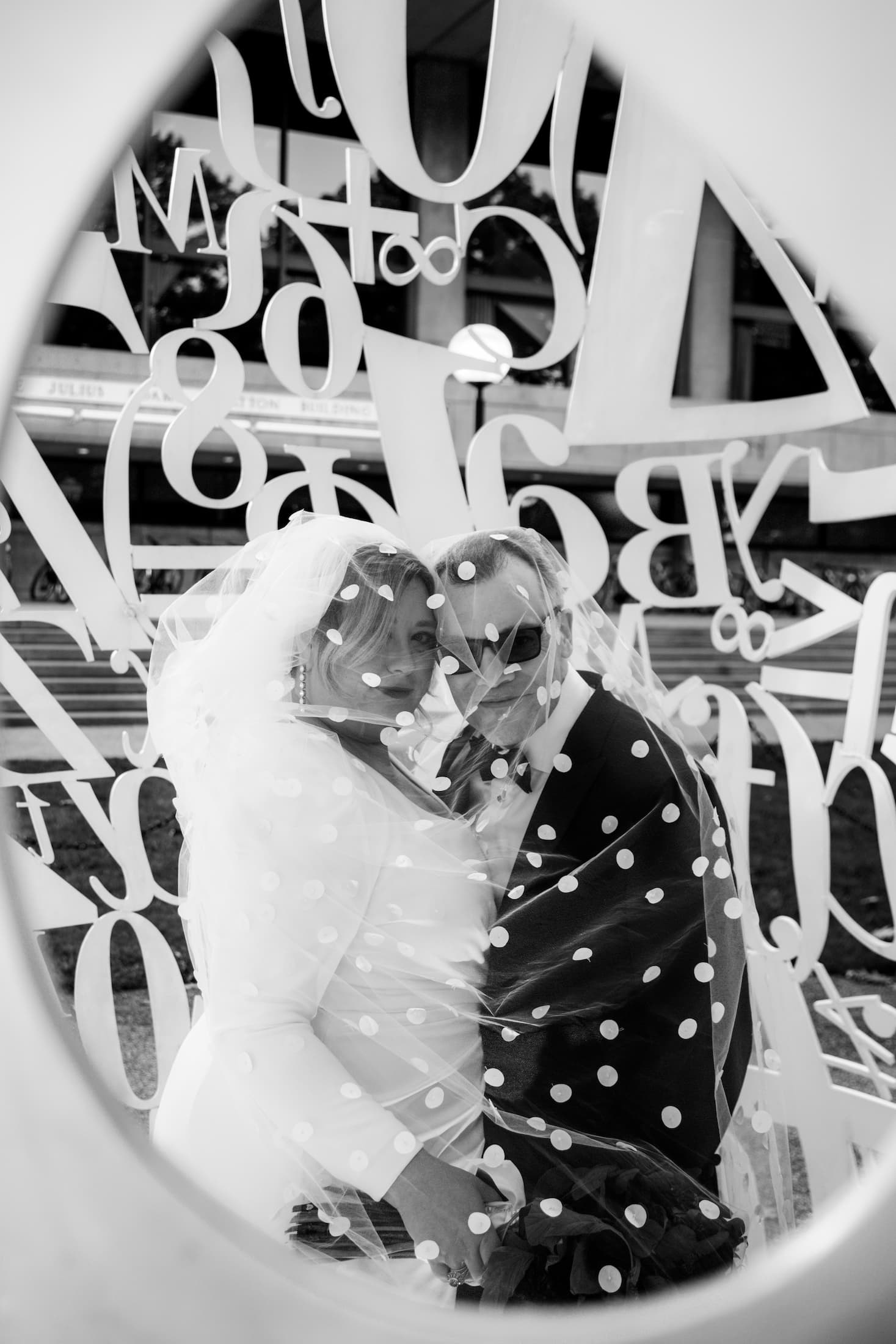 A black and white photo of a bride and a groom wrapped in the bride's polka dotted veil as they stand inside the Alchemist sculpture at MIT in Cambridge, MA - photographed by Spagnolo Photography for their micro-wedding photography package.