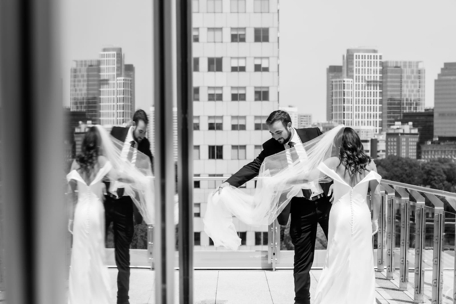 A black and white photograph of a groom helping the bride with her veil that is blowing in the wind - their reflections in the glass window of the MIT Strand Center takes up half the image, which was captured by Boston wedding photographer Spagnolo Photography during their comprehensive Boston wedding photography package that included engagement photography session and an heirloom album.