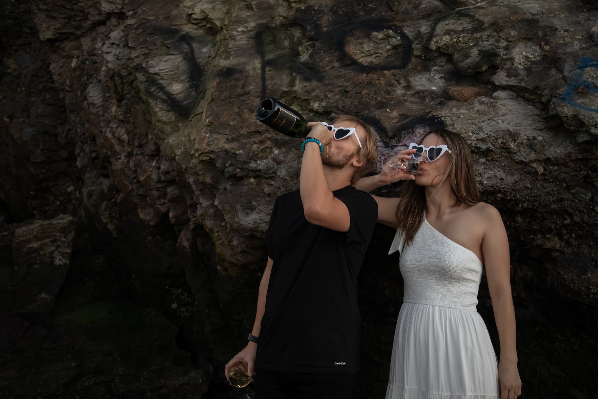 A man dressed in black, and a woman in a white dress, both wearing heart-shaped sunglasses, drink white wine. THe man drinks from the bottle, the woman from a glass, behind them a beachside cliff with graffiti near Boston - photographed by Spagnolo Photography during a Couples Photography session that was part of the couple's wider wedding photography package.