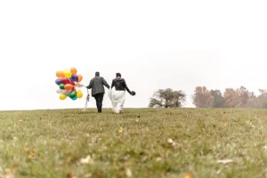An LGBTQ couple walks away from the camera holding rainbow balloons in a field in the rain - the image is featured along with their rave review of their LGBTQ-friendly boston wedding photographer Spagnolo Photography