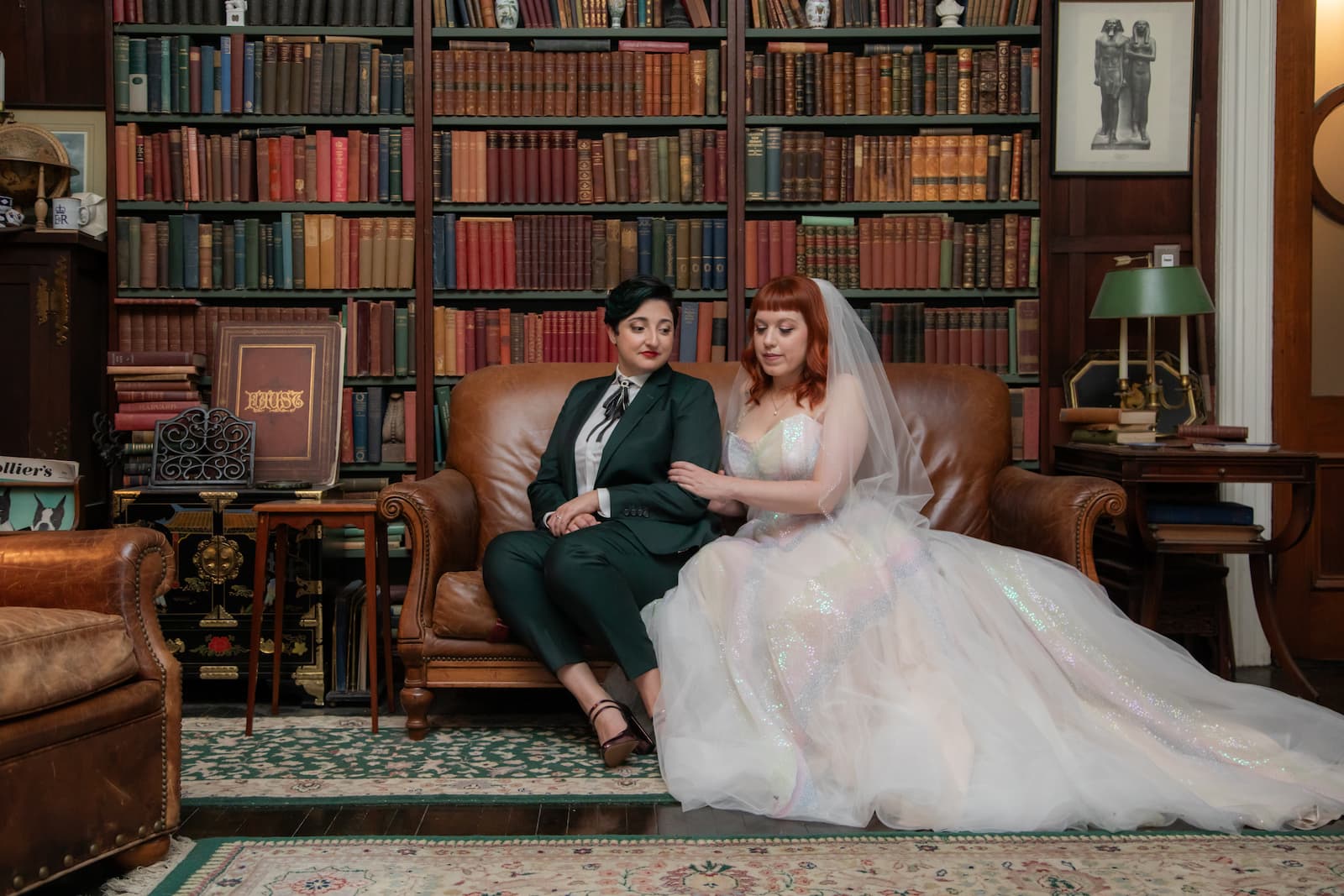 Two brides sit on a brown leather couch surrounded by books and antiques at Diehl Marcus in Salem, MA, before their wedding at The Ledger restaurant, photographed by artistic wedding photographer Spagnolo Photography as part of their wedding photography package that included two engagement sessions and an heirloom album