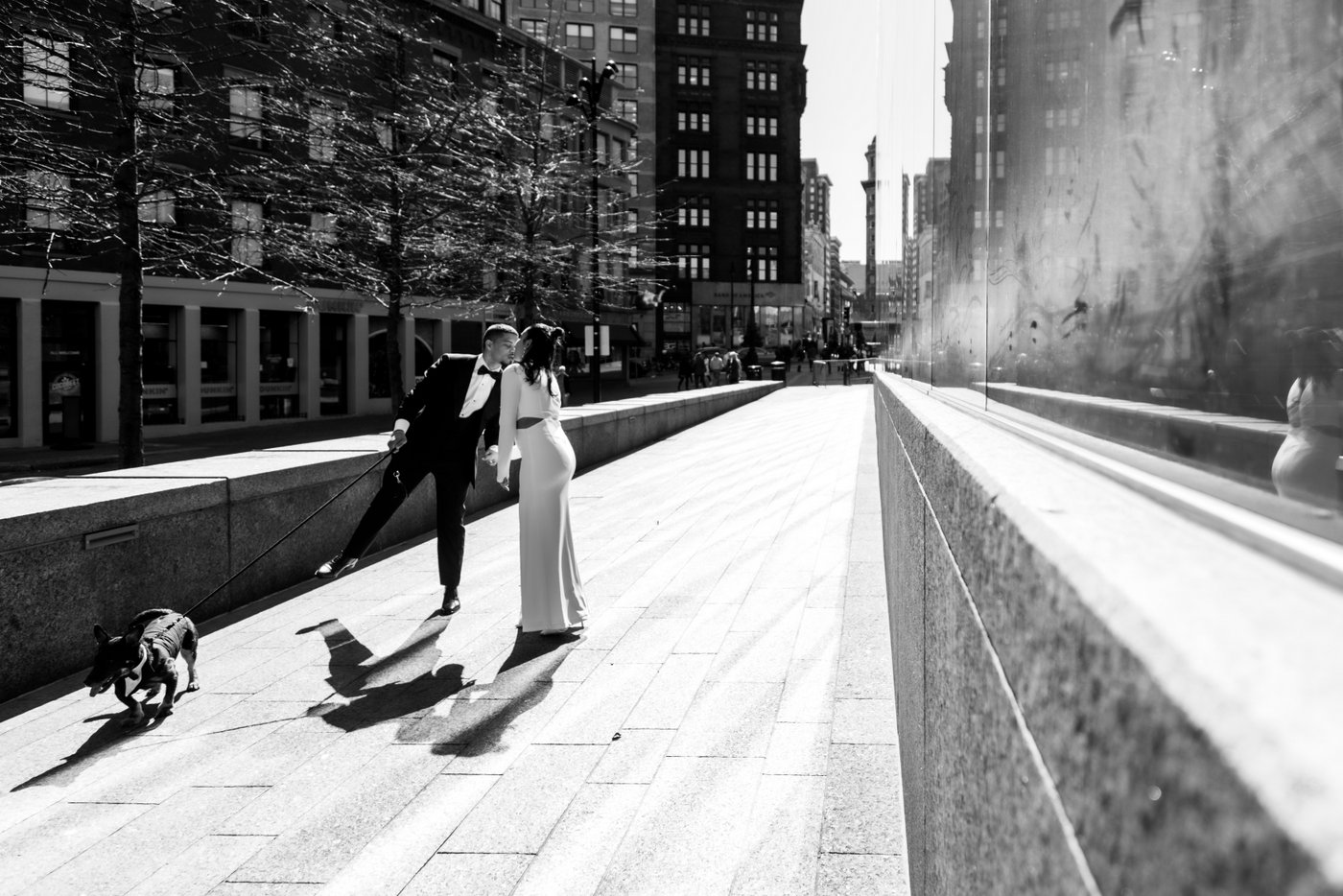 A black and white image of the bride, the groom, and their dog in a tux, walking on the sidewalk towards Boston City Hall where the couple will get married