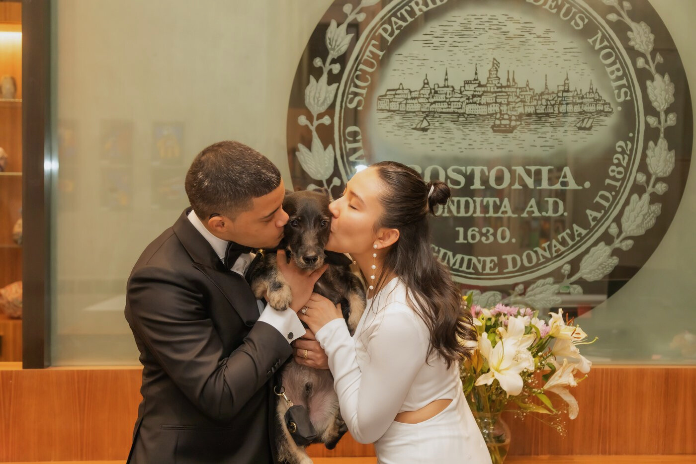The bride and groom kiss their dog by the Boston seal at Boston City Hall on their wedding day
