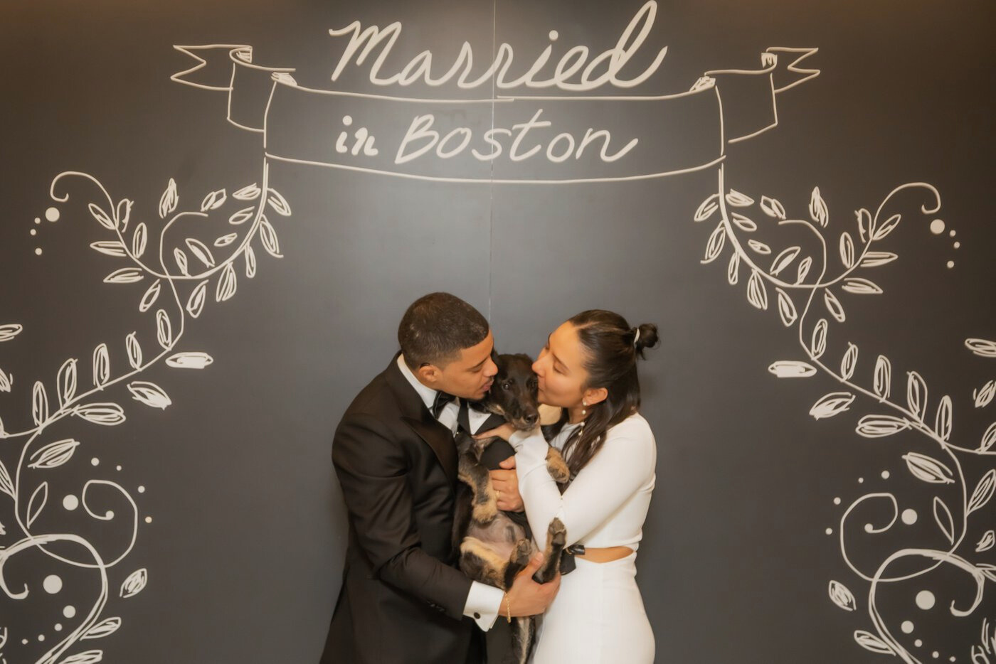 Boston City Hall wedding photographers Spagnolo Photography capturing a bride and a groom kissing their dog under the "Married in Boston" sign following their Boston City Hall elopement.
