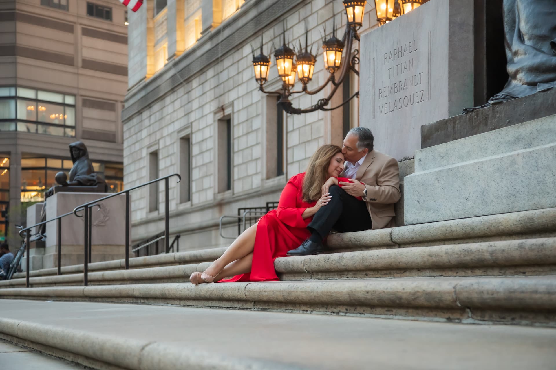 A romantic Boston engagement portrait of a man in a tan suit jacket sitting on the steps of the Boston Public Library and kissing the forehead of a woman in a long red dress who is leaning her head against his knee - photographed at sunset by Boston engagement photographers Spagnolo Photography.