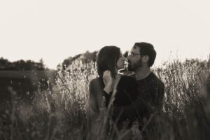 A black and white image of a man and a woman sitting in a field with sunset lighting the tips of the grass around them, photographed by Boston engagement photographers Spagnolo Photography in Andover as part of their wedding photography package - the image is featured on the photographers' "Couples Photography Services & Pricing" page