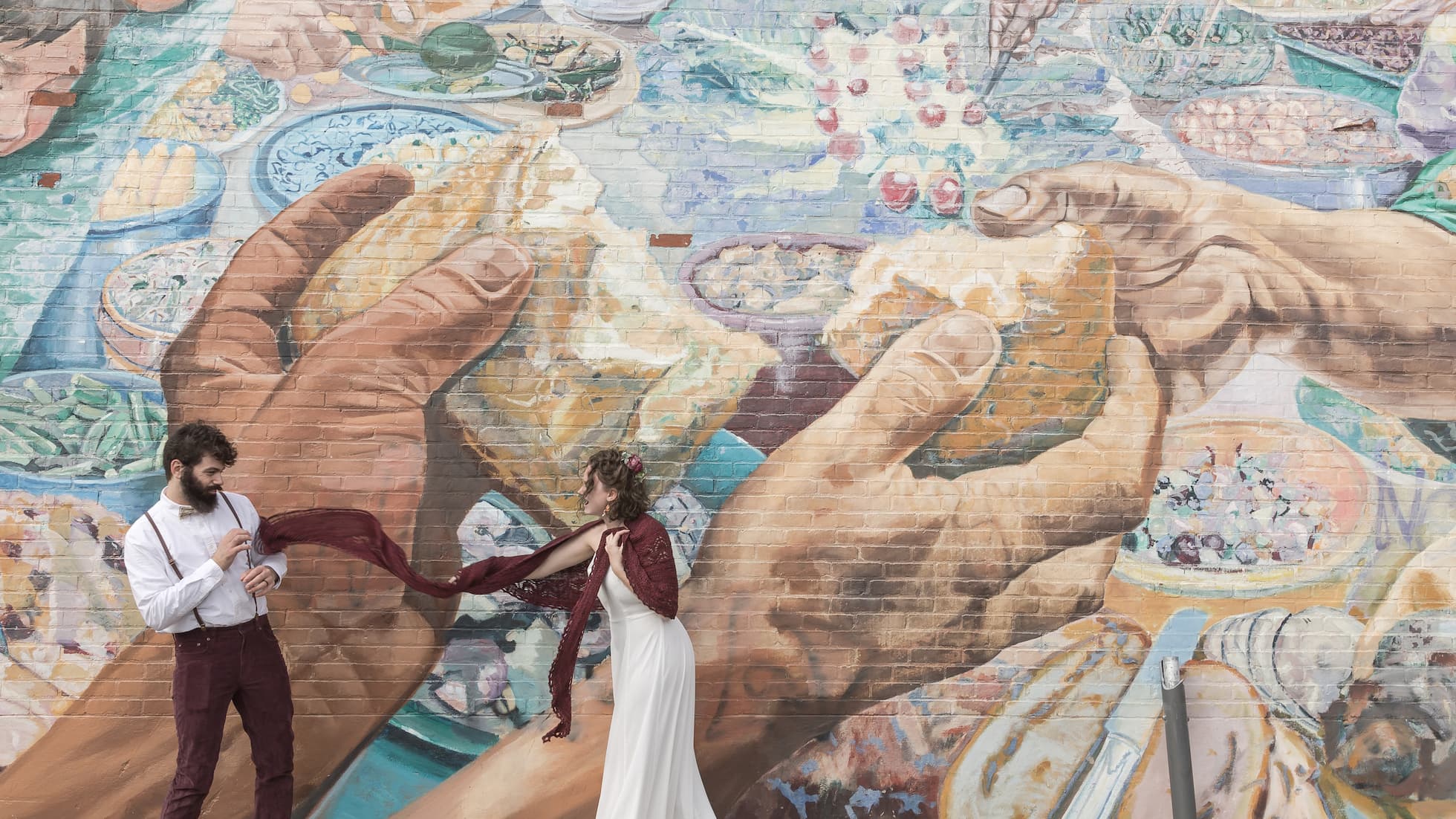Boston wedding photographers Spagnolo Photography capturing a creative wedding elopement moment in front of a large mural depicting hands sharing bread with a bride in a white wedding gown tossing one end of her burgundy red scarf towards the groom who is wearing burgundy pants and suspenders, photographed in Cambridge, MA.