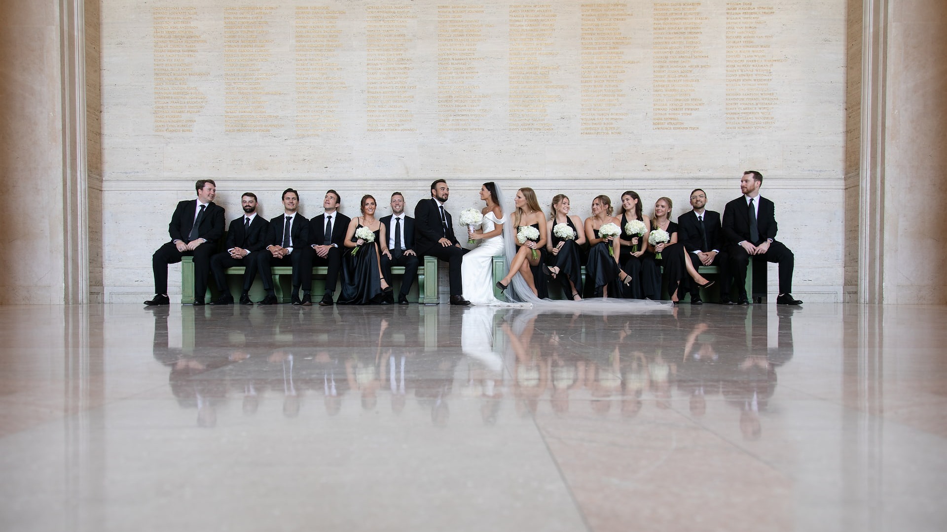 Boston wedding photographers Spagnolo Photography capturing a creative portrait of a bride and a groom and their wedding party sitting on benches in MIT's Lobby 10, with their reflections mirrored on the marble floor, during their MIT wedding.