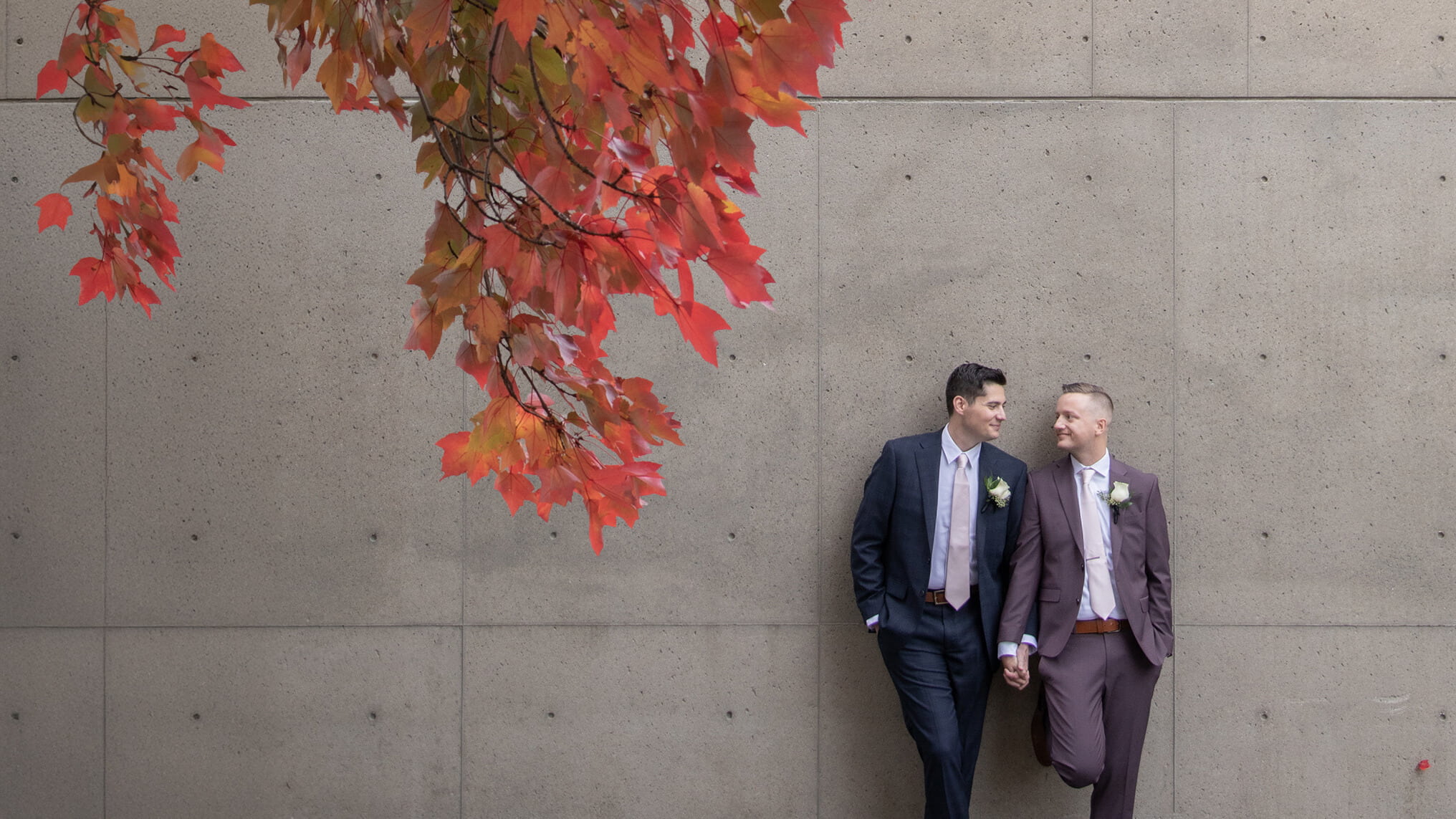 Boston wedding photographers Spagnolo Photography capturing two grooms moments after their City Hall elopement, leaning against a cement wall that is framed by the red leaves of a tree.