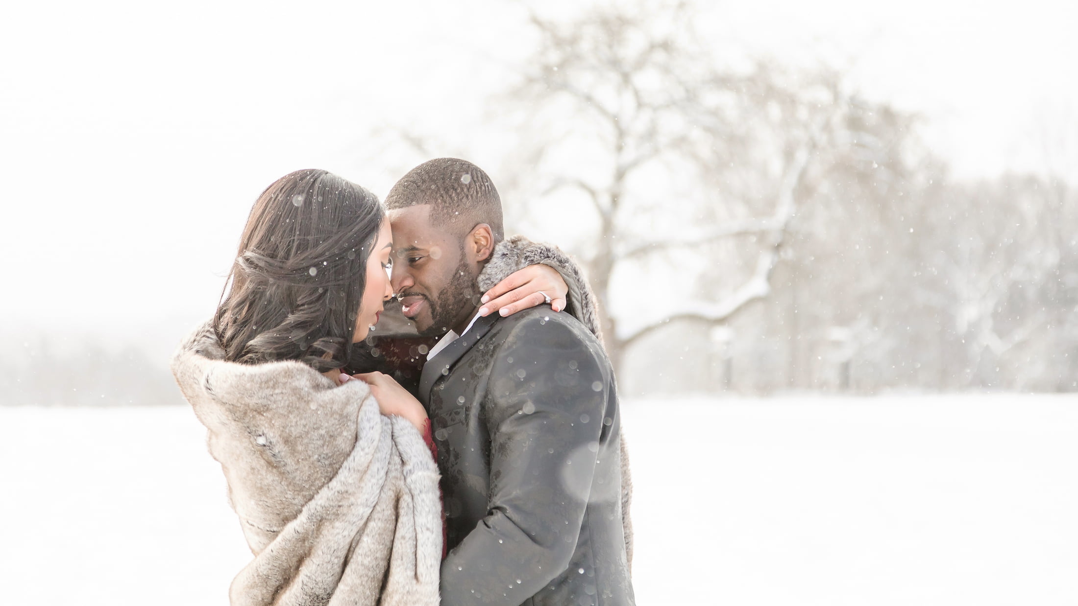 Boston wedding photographers Spagnolo photography capturing a Massachusetts winter engagement photo that shows a man and a woman wrapped in a cozy fur blanket during a snowstorm in a snow-covered field.