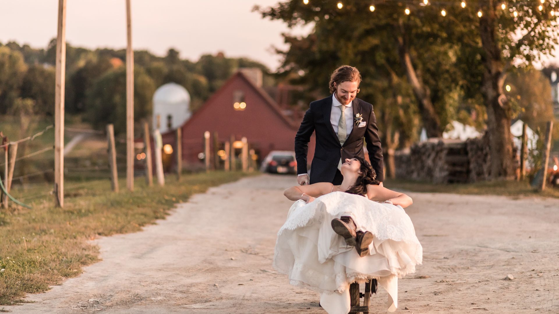 Boston wedding photographers Spagnolo Photography capturing a creative wedding portrait of a bride sitting in a wheelbarrow that the groom pushes in front of a red barn under string lights at a Gibbet Hill wedding in Groton, MA.