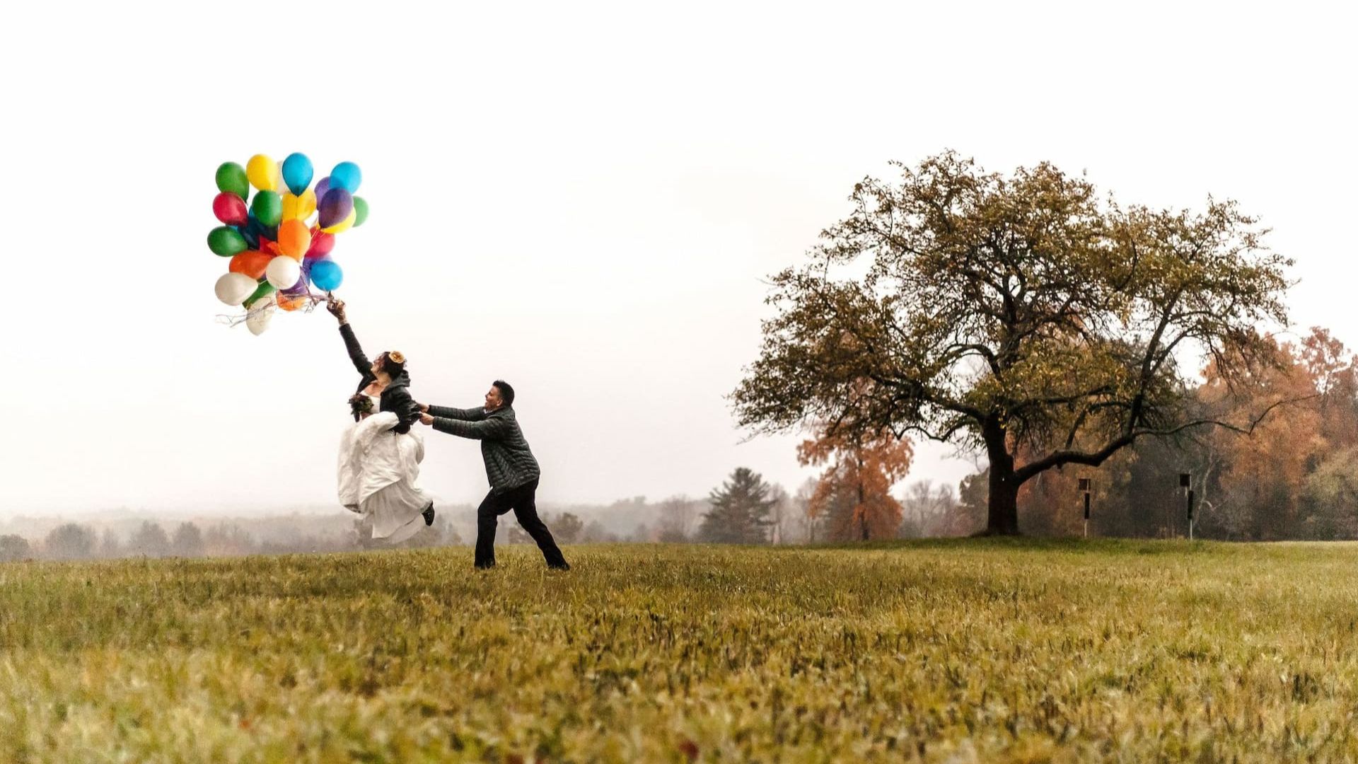 Boston wedding photographers Spagnolo Photography capturing a creative, artistic, and candid LGBTQ elopement photo of a couple running and jumping in a field under pouring rain while holding a vibrant colorful bunch of balloons, which makes them look like they are about to fly.