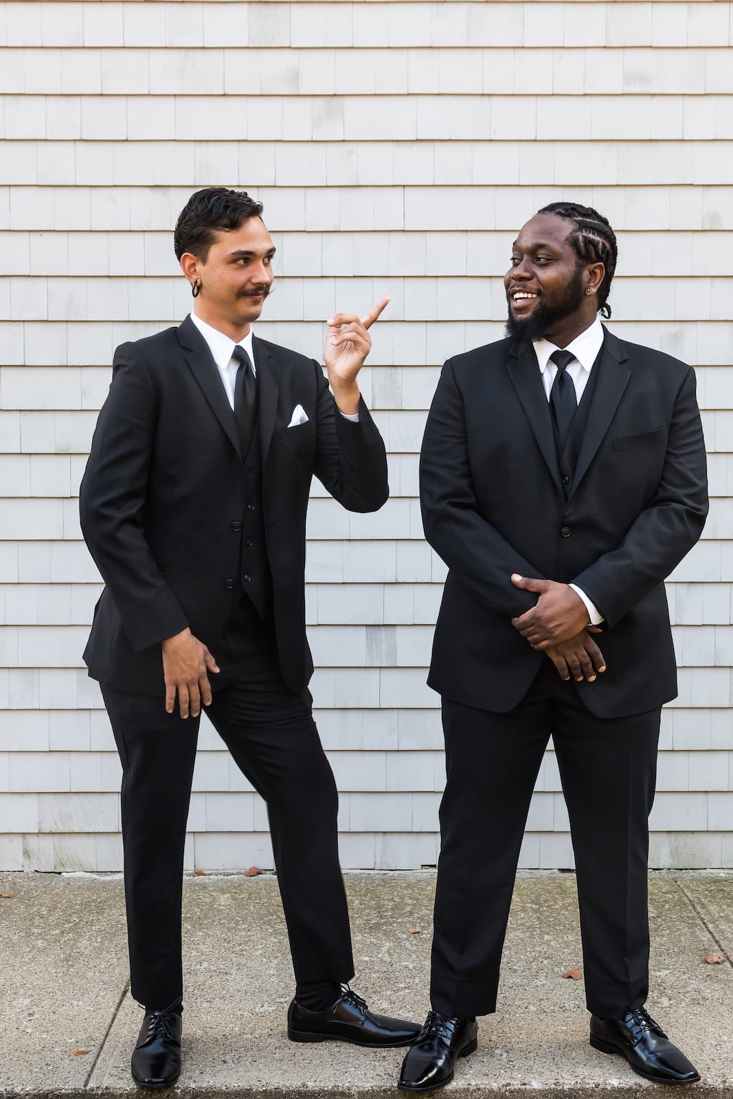 Two groomsmen stand in front of a shingled blue-gray wall, chatting, with one pointing up as the other laughs - a funny candid photo from a Gloucester wedding by Massachusetts wedding photographers Aaron and Nanore of Spagnolo Photography