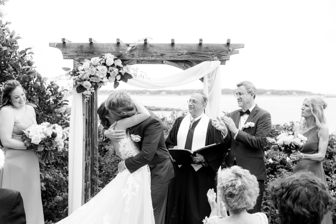 A black and white image of a bride and groom's first kiss at the altar during their wedding ceremony on the oceanside lawn of Hammond Castle