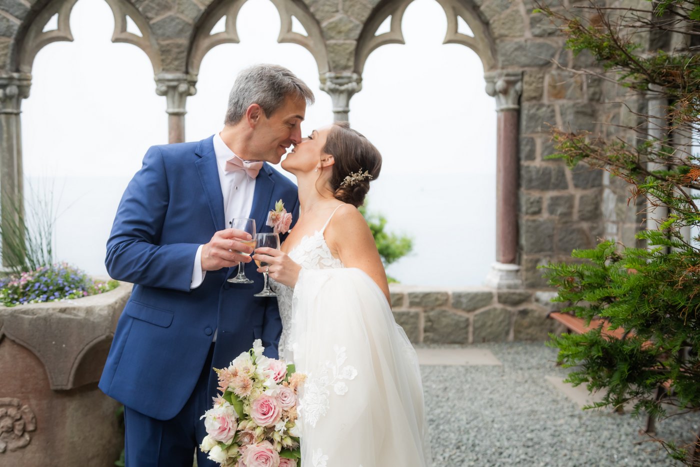 The bride and groom kiss while toasting their champagne glasses by Hammond Castle's stone arches following their wedding ceremony