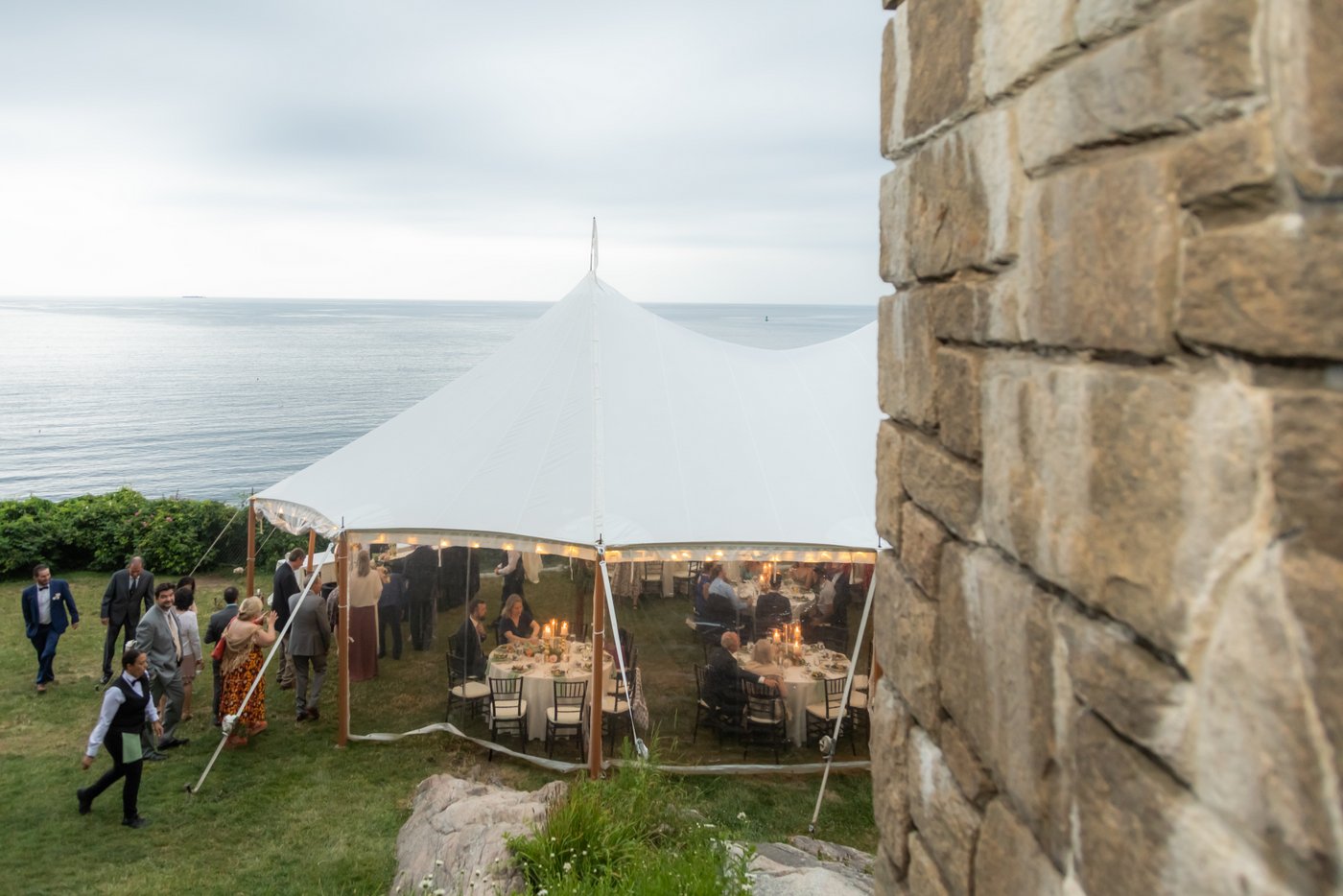 Guests beginning to enter the wedding reception tent with string lights on Hammond Castle's Oceanside lawn overlooking the water, photographed from the top of the back stairs of the castle