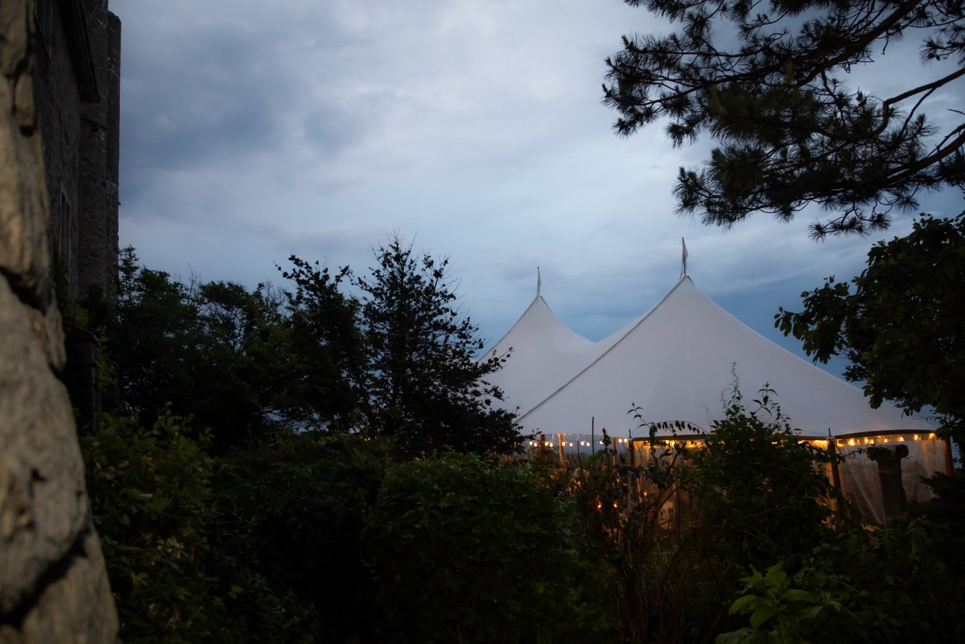 A photograph of the outside of the wedding reception tent on the oceanside lawn of Hammond Castle at dusk with the string lights glowing