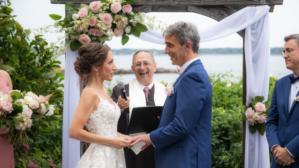 Hammond Castle wedding ceremony portrait showing the bride and groom gazing at each other and smiling as the officiant grins under the floral altar on the oceanside lawn.