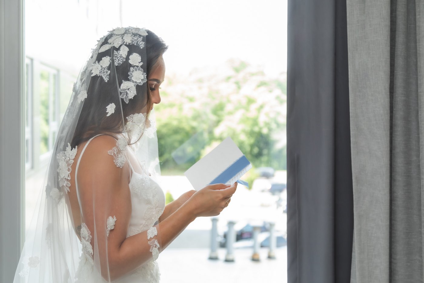 The bride, wearing her wedding gown and lace veil, stands by the window and reads a letter from the groom