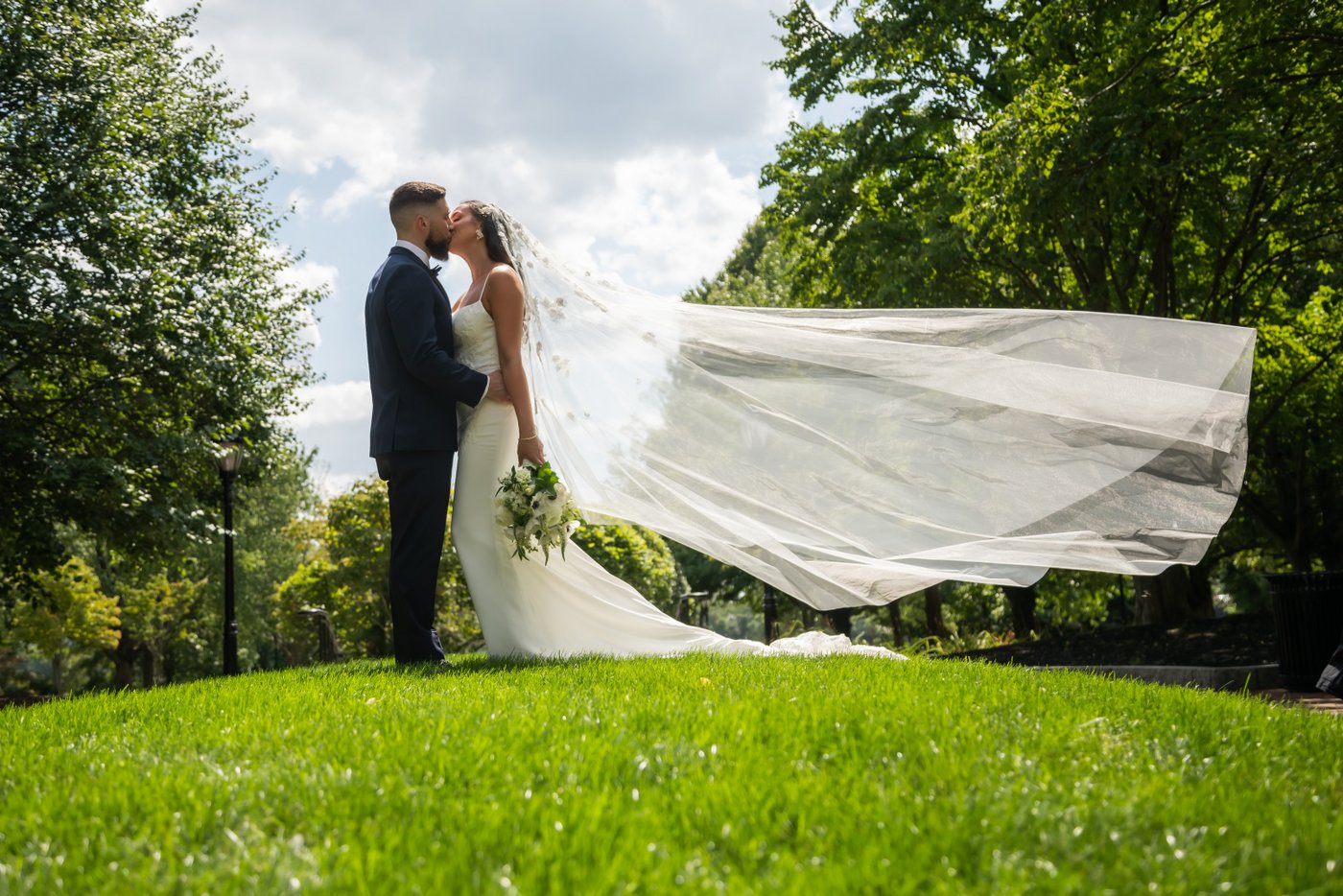 The bride's veil floats in the air as the bride and groom kiss surrounded by greenery and a blue sky
