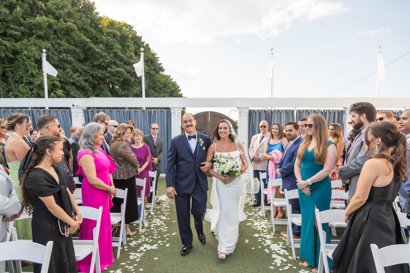 The bride and her father smile as they walk down the aisle overlooking the water at an Oceanview of Nahant wedding in coastal Massachusetts.
