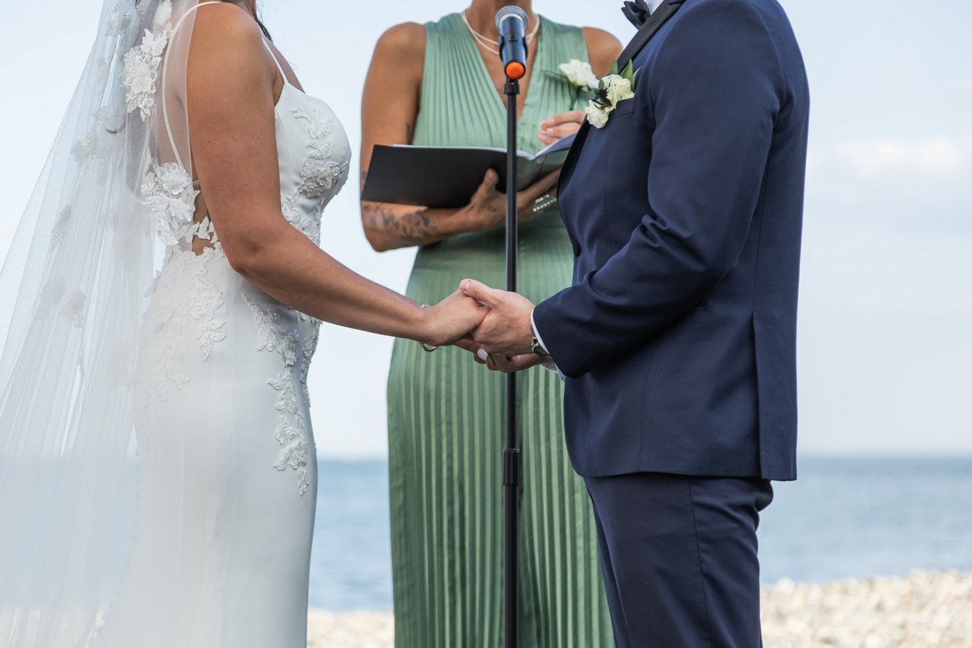 A close-up of the bride and groom's hands holding each other during the Oceanview of Nahant wedding ceremony with the blue ocean behind them