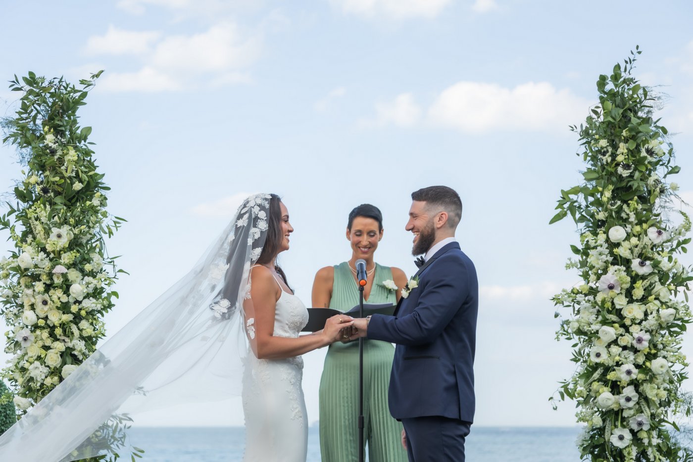 A beautiful photo of the bride and groom exchange rings as the bride's veil floats with the wind during their Oceanview of Nahant wedding