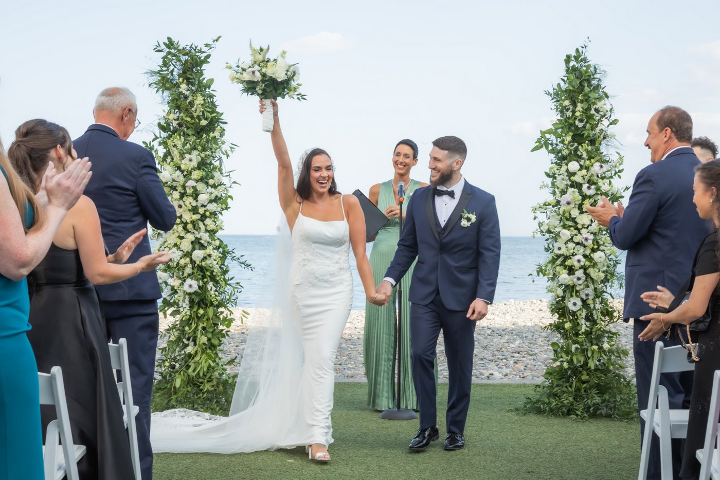 The bride raises her bouquet in the air as she and the groom begin walking back up the aisle holding hands as guests stand and clap during an Oceanview of Nahant wedding by the water
